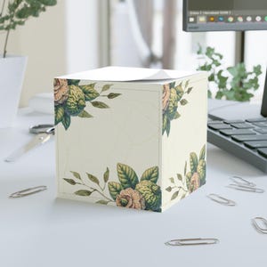 Puede incluir: Un bloc de notas cuadrado con un diseño floral con flores y hojas melocotón y verdes sobre un fondo crema. El bloc de notas está sobre un escritorio blanco con clips y un portátil en el fondo.