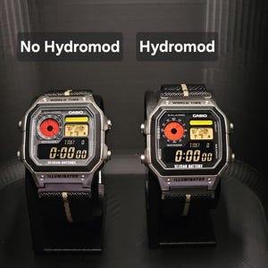 May include: Two digital Casio watches side-by-side, one labeled "No Hydromod" and the other "Hydromod." Both watches have a silver case, black band with yellow stripe, and a digital display showing "0:00 00." The text "ILLUMINATOR" is visible.