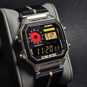 Könnte beinhalten: Eine silberne Casio Digitaluhr mit schwarzem Zifferblatt und einem schwarzen und beigen Nylonarmband. Die Uhr zeigt die Uhrzeit, das Datum und Weltzeitfunktionen an. Die Uhr trägt die Aufschrift "Casio", "World Time" und "Illuminator".