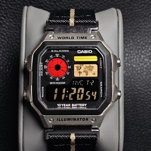May include: A silver digital watch with a black face and a black and beige striped band. The watch face displays the time, world time, and other functions. The watch is labeled "CASIO" and "ILLUMINATOR".