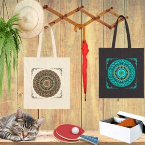 May include: Two tote bags with mandala designs hang on a wooden rack. One is off-white with a brown and green mandala, the other is black with a teal mandala. A red umbrella hangs nearby.