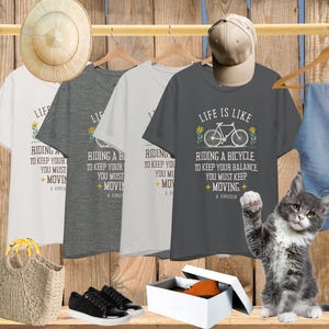 May include: Four short-sleeve t-shirts in white, dark gray, light gray, and charcoal gray, each featuring a graphic of a bicycle, yellow flowers, and the text: Life is like riding a bicycle. To keep your balance, you must keep moving. A. Einstein.