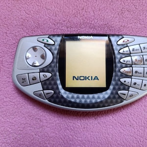May include: A silver and black Nokia mobile phone with a horizontal, oval-shaped body. The device features a central screen displaying the Nokia logo, a directional pad on the left, and a standard numeric keypad on the right with call control buttons.