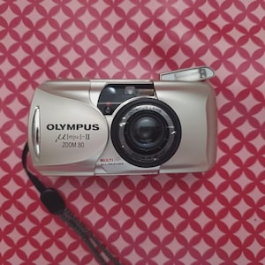May include: A silver Olympus mju: II Zoom 80 point-and-shoot film camera with a black lens barrel, a built-in flash, and a black wrist strap. The front of the camera features the text OLYMPUS, mju: II, ZOOM 80, and MULTI ALL-WEATHER printed in black.