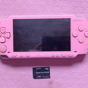 Può includere: Una console di gioco portatile Sony PSP rosa con schermo nero, pad direzionale e pulsanti di azione. Sotto la console si trova una scheda di memoria nera Sony Memory Stick PRO Duo da 2 GB con il logo MagicGate stampato sul davanti.