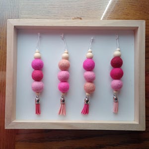 May include: Four decorative hanging ornaments with a wooden frame. Each ornament features a string, wooden bead, and a series of felt balls in shades of pink and red, topped with a small tassel. The ornaments are displayed against a white background.