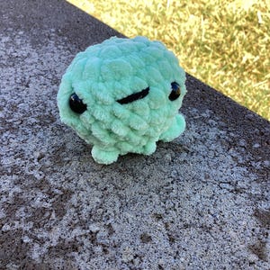 May include: A mint green crocheted octopus plush toy with black eyes and a black stitched mouth. The plush is sitting on a gray concrete surface with a blurred green background.