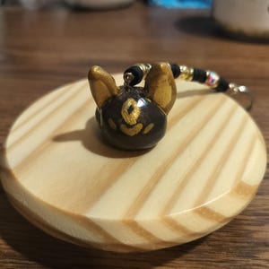 Puede incluir: Un charm hecho a mano con la forma de la cabeza de un perro, con orejas y detalles dorados. El charm es de color marrón oscuro con detalles dorados y está unido a una cadena de cuentas negras y doradas. Descansa sobre una base de madera redonda y clara.