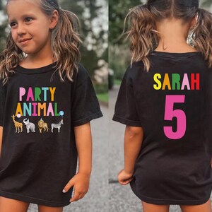 May include: Black t-shirt with the words "PARTY ANIMAL" in colorful letters and animal graphics. The back of the shirt has the name "SARAH" and the number "5" in bright colors. The shirt is worn by a young girl.