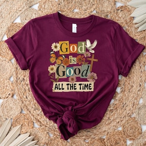 May include: Burgundy t-shirt with the text "God is Good ALL THE TIME" in a colorful, floral design. The design includes butterflies, flowers, a dove, and a cross. The shirt is knotted at the bottom.