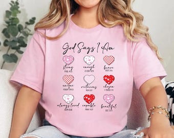 May include: A light pink t-shirt featuring the text God Says I Am above nine heart icons. Each heart includes a descriptive word and a Bible verse reference, including strong, enough, brave, unique, victorious, chosen, loved, capable, and beautiful.