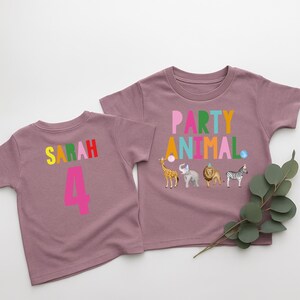 May include: Two mauve-colored toddler t-shirts. One shirt has the name "Sarah" and the number "4" in colorful letters. The other shirt says "Party Animal" with animal graphics, including a giraffe, elephant, lion, and zebra.