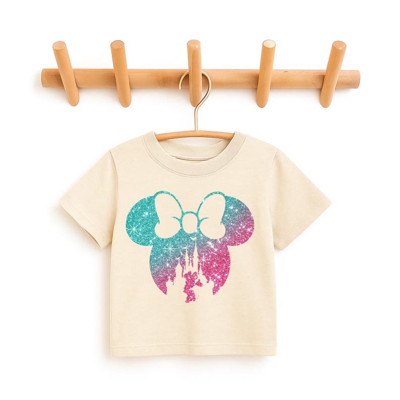 Puede incluir: Una camiseta color crema con una silueta brillante de Minnie Mouse en verde azulado y rosa. El dise&ntilde;o incluye un lazo y una silueta de castillo. La camiseta est&aacute; colgada de una percha de madera sujeta a un perchero de madera.