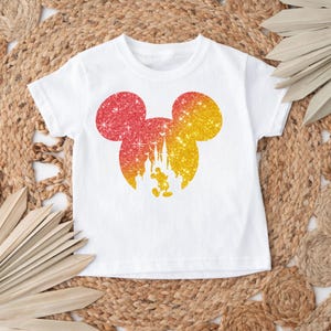 Puede incluir: Camiseta blanca con un dise&ntilde;o brillante de Mickey Mouse en rojo y amarillo. El dise&ntilde;o incluye un castillo y Mickey Mouse. La camiseta es de cuello redondo y manga corta.
