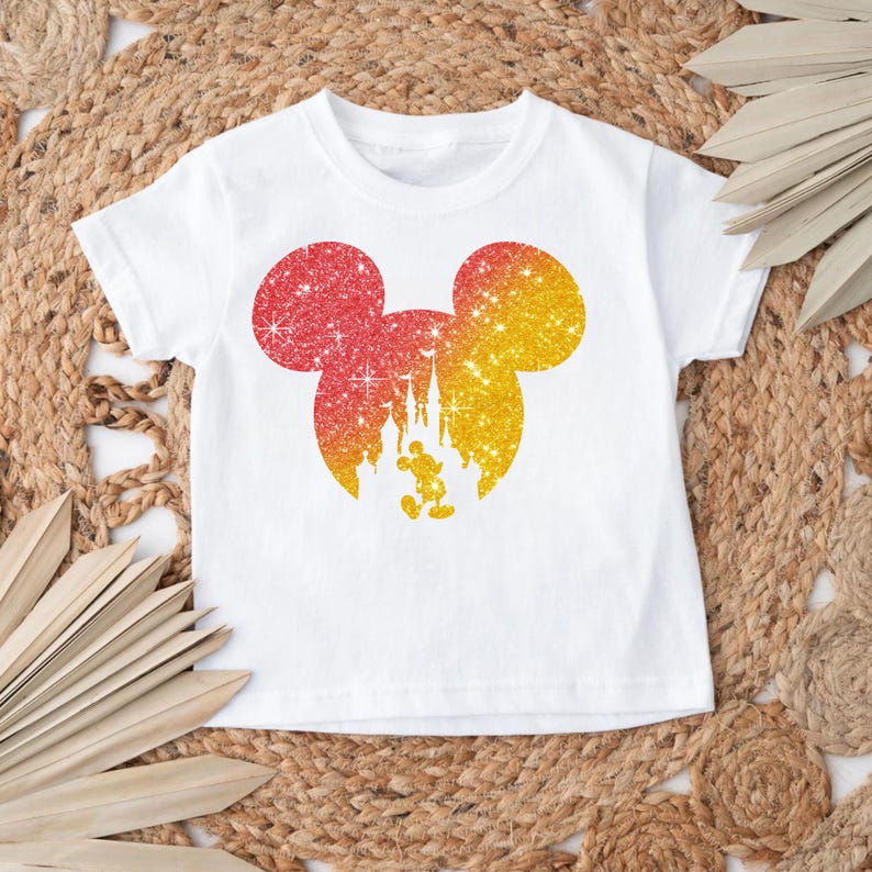 Puede incluir: Camiseta blanca con un dise&ntilde;o brillante de Mickey Mouse en rojo y amarillo. El dise&ntilde;o incluye un castillo y Mickey Mouse. La camiseta es de cuello redondo y manga corta.