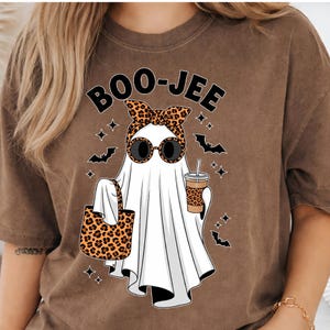 May include: A brown short-sleeve t-shirt featuring a graphic of a white ghost wearing leopard print sunglasses and a matching headband. The ghost holds a leopard print tote bag and a coffee cup. The text BOO-JEE is printed above the ghost.