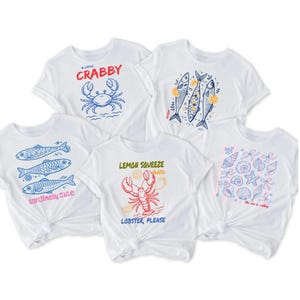 May include: White t-shirts with various sea-themed designs. One shirt features a blue crab with the text "A Little Crabby." Another displays three fish with the words "sardinely cute." Other designs include a lobster, fish, and seashells.