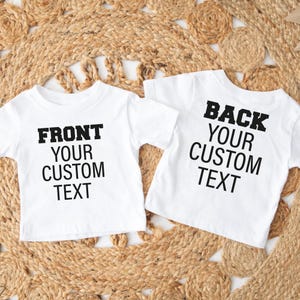 May include: Two white t-shirts with black text. One shirt reads "FRONT YOUR CUSTOM TEXT", and the other reads "BACK YOUR CUSTOM TEXT". The shirts are short-sleeved and appear to be for children. They are displayed on a woven, beige surface.