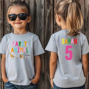 May include: A gray t-shirt with the words "PARTY ANIMAL" in colorful letters and animal illustrations. The back of the shirt has the name "SARAH" and the number "5" in bright colors. The shirt is worn by a young child.