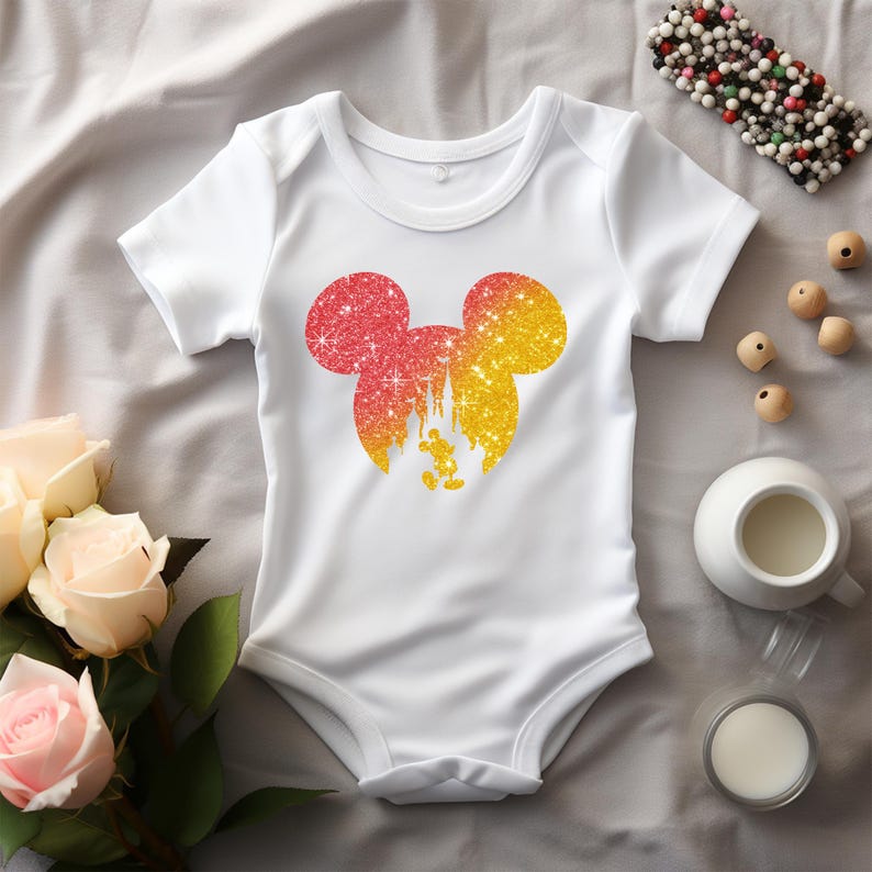 Puede incluir: Body de beb&eacute; blanco con un dise&ntilde;o de silueta de Mickey Mouse brillante en tonos rojos y amarillos. El dise&ntilde;o incluye un castillo y Mickey Mouse. El body est&aacute; rodeado de rosas, una peque&ntilde;a taza de leche y un dulce de chocolate.