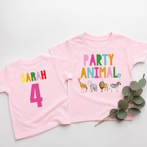 May include: Two pink toddler t-shirts. One shirt has the name "SARAH" in rainbow colors and the number "4" in pink. The other shirt says "PARTY ANIMAL" in rainbow colors with animal illustrations.