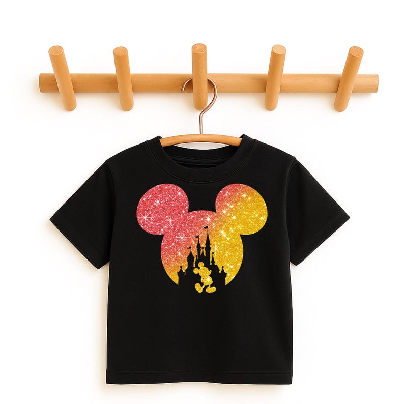 Puede incluir: Camiseta negra con un dise&ntilde;o de cabeza de Mickey Mouse brillante y una silueta de castillo. La cabeza de Mickey Mouse hace la transici&oacute;n de rojo a amarillo. La camiseta est&aacute; colgada en una percha de madera.