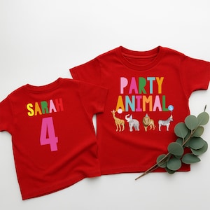 May include: Two red t-shirts. One shirt has the name "Sarah" and the number "4" in colorful letters. The other shirt says "PARTY ANIMAL" with animal illustrations, including a giraffe, elephant, lion, and zebra.