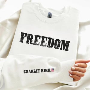 May include: A white sweatshirt with the word "FREEDOM" printed in large, distressed black letters across the chest. The sleeve features the text "CHARLIE KIRK" with a small American flag graphic.
