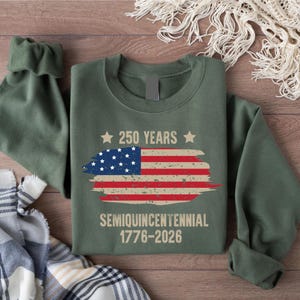 May include: An olive green sweatshirt featuring a distressed American flag design with the text "250 YEARS" above and "SEMIQUINCENTENNIAL 1776-2026" below. The sweatshirt is folded, with the sleeves visible.