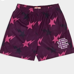 Könnte beinhalten: Lilafarbene Basketballshorts mit einem pinkfarbenen und schwarzen Sternmuster und einem weißen "BE"-Logo am rechten Bein.