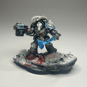May include: A black and silver painted miniature space marine with a blue energy sword and a skull helmet. The miniature is standing on a snowy base with red blood splatter.