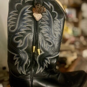 May include: Black leather cowboy boot with intricate blue and white stitching. A decorative feather and stone accent hangs from the boot's pull strap. Gold-colored metal accents are also present.