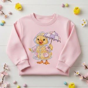 Pode incluir: Um moletom rosa claro com um desenho de um pintinho de desenho animado usando uma capa de chuva e segurando um guarda-chuva xadrez roxo e branco. O pintinho é amarelo com bico e pés laranja. O moletom tem mangas compridas e gola redonda.