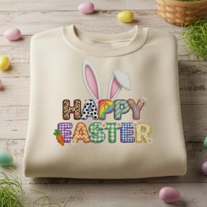 Pode incluir: Camisola creme com um gráfico colorido "Feliz Páscoa". O design apresenta orelhas de coelho e letras com vários padrões, incluindo estampa de leopardo, arco-íris e xadrez. Um gráfico de cenoura também está incluído. Roupas com tema de Páscoa.
