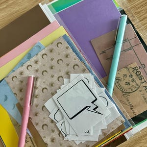 May include: A collection of stationery items, including colourful paper, stickers, and pens. The image features a light pink pen, a light blue pen, and speech bubble stickers. Various paper colours include brown, pink, yellow, and purple.