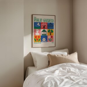 Op de afbeelding: Een ingelijste poster getiteld "The Public Gardens" met kleurrijke vierkante illustraties. Het kunstwerk toont een vogel, boom, poort, buste, tuinhuisje, bloemen en het woord "Halifax". De poster wordt weergegeven boven een bed met wit beddengoed.
