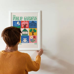 Op de afbeelding: Een ingelijste kunstprint getiteld "The Public Gardens" met kleurrijke illustraties van een eend, boom, poort, prieel, fontein en bloemen. De persoon op de foto draagt een gele trui.