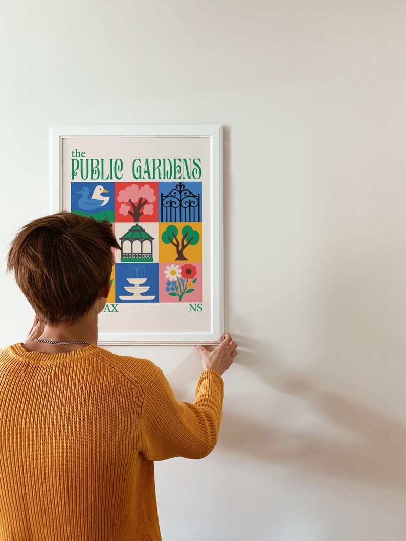 Op de afbeelding: Een ingelijste kunstprint getiteld "The Public Gardens" met kleurrijke illustraties van een eend, boom, poort, prieel, fontein en bloemen. De persoon op de foto draagt een gele trui.