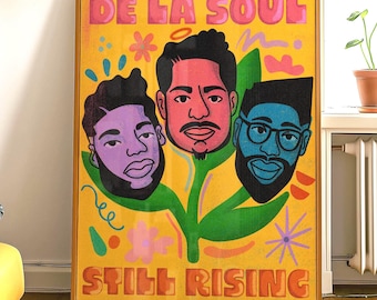 Op de afbeelding: Een ingelijste poster met de tekst "DE LA SOUL" in roze en "STILL RISING" in oranje. De afbeelding toont drie gestileerde gezichten in paars, rood en blauw, tegen een gele achtergrond met bloemaccenten. De lijst is warmbruin.