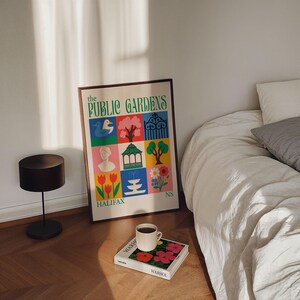 Op de afbeelding: Een ingelijste poster met de tekst "The Public Gardens" en kleurrijke illustraties van parkelementen. Een koffiekopje staat op een boek met bloemenkunst. Een zwarte lamp en een bed staan ook in de afbeelding.