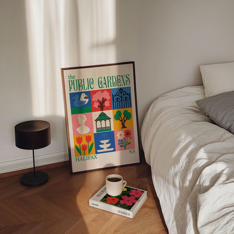 Op de afbeelding: Een ingelijste poster met de tekst "The Public Gardens" en kleurrijke illustraties van parkelementen. Een koffiekopje staat op een boek met bloemenkunst. Een zwarte lamp en een bed staan ook in de afbeelding.