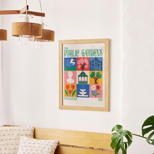 Op de afbeelding: Een ingelijste poster met de tekst "The Public Gardens" en kleurrijke illustraties van een zwaan, bomen en bloemen. De poster zit in een licht houten frame en hangt aan een witte muur. Een houten bank en een hanglamp zijn ook zichtbaar.