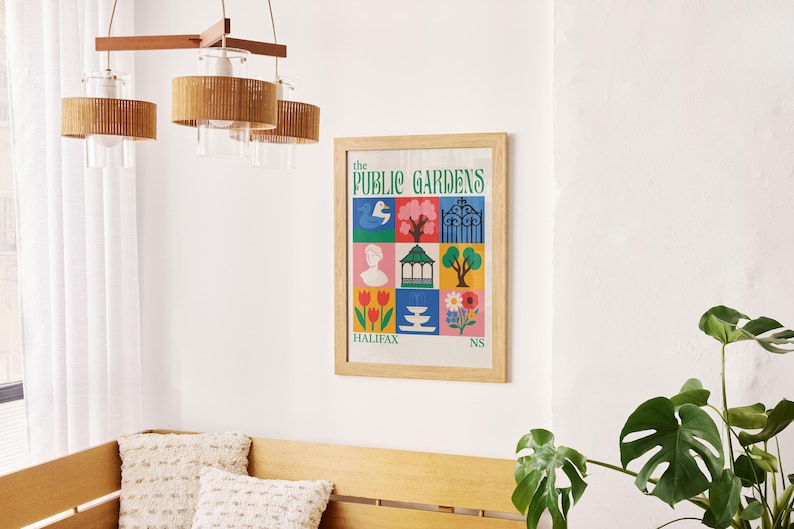 Op de afbeelding: Een ingelijste poster met de tekst "The Public Gardens" en kleurrijke illustraties van een zwaan, bomen en bloemen. De poster zit in een licht houten frame en hangt aan een witte muur. Een houten bank en een hanglamp zijn ook zichtbaar.