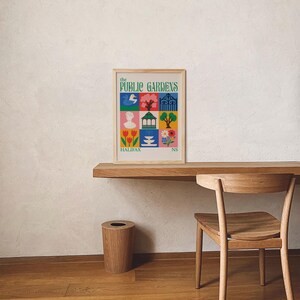 Op de afbeelding: Een ingelijste poster met de tekst "the PUBLIC GARDENS" en "HALIFAX NS" in een kleurrijk rasterontwerp. De poster staat op een houten bureau met een houten stoel en een houten prullenbak. Het ontwerp van de poster bevat illustraties van een vogel, bloemen en een standbeeld.