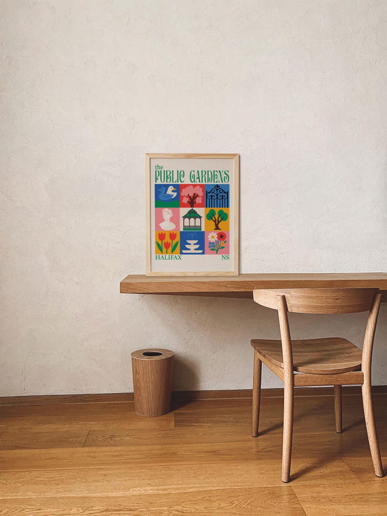 Op de afbeelding: Een ingelijste poster met de tekst "the PUBLIC GARDENS" en "HALIFAX NS" in een kleurrijk rasterontwerp. De poster staat op een houten bureau met een houten stoel en een houten prullenbak. Het ontwerp van de poster bevat illustraties van een vogel, bloemen en een standbeeld.