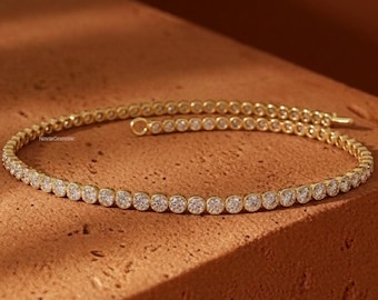 May include: A delicate gold bracelet with a continuous row of round, clear gemstones. The bracelet is set against a warm, brown background, highlighting its sparkle. The bracelet is a tennis style, with a simple clasp.