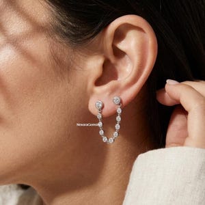 Op de afbeelding: Zilveren oorbellen met een ketting van kleine, ronde, heldere edelstenen. De oorbellen zijn verbonden door een delicate ketting van kleinere stenen, waardoor een watervaleffect ontstaat. De oorbellen worden op het oor gedragen.