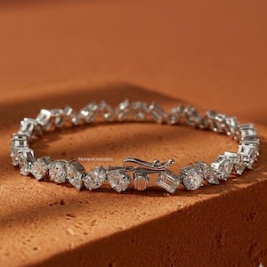Puede incluir: Una pulsera plateada con múltiples piedras preciosas transparentes de varias formas, incluyendo cortes redondos, de pera y esmeralda. La pulsera se asegura con un cierre y se encuentra sobre una superficie marrón texturizada. La marca "NovaraGemsInc" es visible.