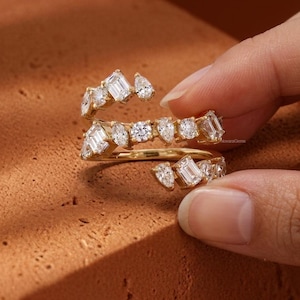 May include: A gold bypass ring featuring a spiral design adorned with multiple clear, faceted stones in various cuts, including emerald, pear, and round shapes, arranged along the band to create a sparkling, multi-layered appearance.