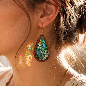 Puede incluir: Un pendiente en forma de lágrima con un marco de madera oscura que rodea una pieza de estilo vitral colorido. La pieza muestra un vibrante patrón floral en tonos verdes, azules, naranjas y blancos, suspendido por un gancho de metal dorado.